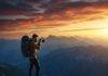 Best Hiking Trails for Stunning Views and Photography Lovers A hiker on a cliff edge holds a camera, capturing the vast mountain range under a dramatic sunset sky filled with warm orange