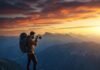 Best Hiking Trails for Stunning Views and Photography Lovers A hiker on a cliff edge holds a camera, capturing the vast mountain range under a dramatic sunset sky filled with warm orange